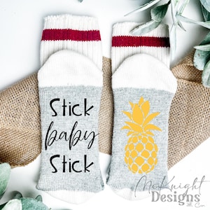Könnte beinhalten: Ein Paar weiße und graue Socken mit roten Streifen. Eine Socke hat den Text "Stick baby Stick" aufgedruckt. Die andere Socke hat ein gelbes Ananas-Design.