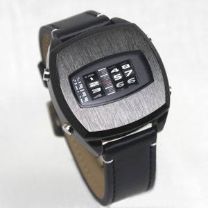 Könnte beinhalten: Eine schwarze Armbanduhr mit einem gebürsteten Metallzifferblatt und einem schwarzen Lederarmband. Das Zifferblatt der Uhr hat eine digitale Anzeige mit den Zahlen 1 bis 8.