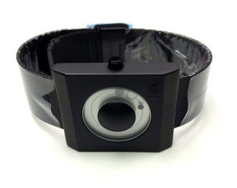 May include: A black square watch with a black band. The watch face has a circular design with a smaller circle in the center. The watch has a small button on the top.
