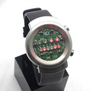 May include: A silver wristwatch with a black leather strap. The watch face is a green circuit board with red LED lights that display the time. The watch face says "HOUR" and "MINUTE".