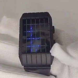 May include: A black wristwatch with a square face and a grid-like display. The display shows the time in blue LED lights.