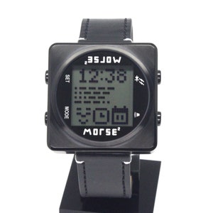 Może przedstawiać: Czarny, cyfrowy zegarek na rękę z kwadratową tarczą i czarnym skórzanym paskiem. Tarcza zegarka wyświetla godzinę, datę i symbol kodu Morse'a. Na tarczy zegarka znajduje się również napis "MORSE".