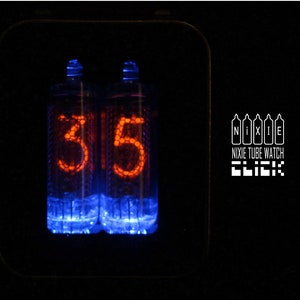 Könnte beinhalten: Nahaufnahme einer Nixie-Röhrenuhr, die die Zeit 3:15 anzeigt. Das Zifferblatt der Uhr ist mit einem blauen Schein beleuchtet, und die Zahlen sind orange. Die Uhr ist mit "Nixie Tube Watch" und "Click" beschriftet.