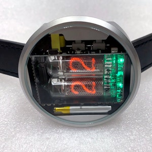 Puede incluir: Un reloj redondo plateado con una correa de cuero negro. La esfera del reloj es transparente y muestra dos tubos Nixie rojos que muestran el número 22 y un tubo Nixie verde que muestra el texto "NixieTube IN-16".