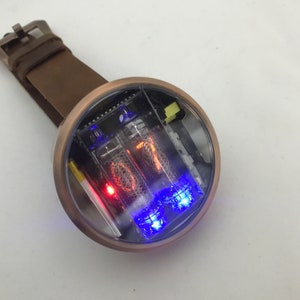 Può includere: Un orologio rotondo di colore bronzo con un quadrante trasparente e un cinturino in pelle marrone. Il quadrante dell'orologio presenta un display digitale con luci rosse e blu.
