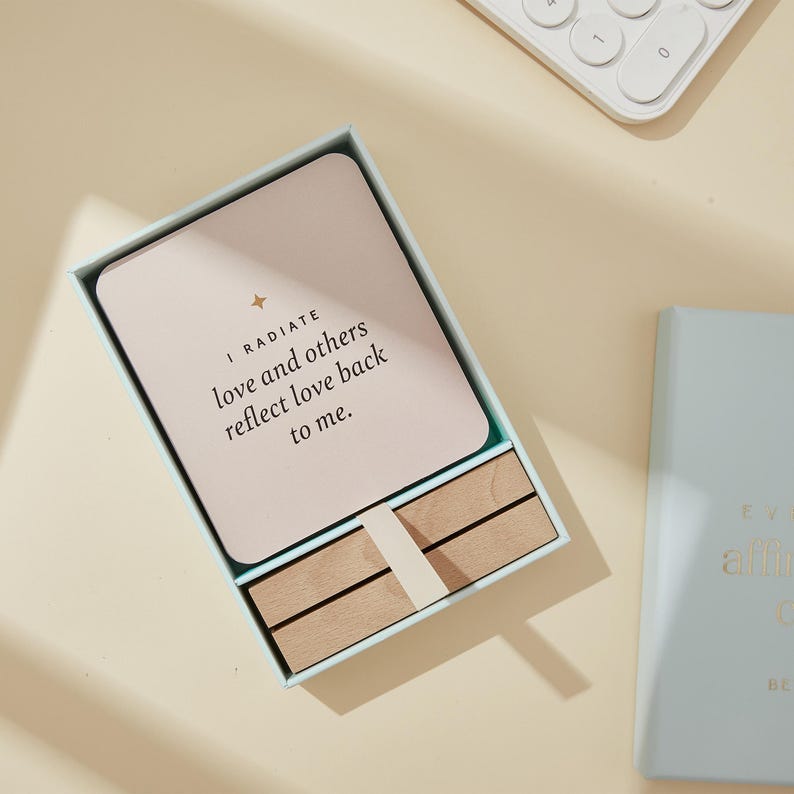 May include: A light blue box containing affirmation cards. The cards are light pink with the text "I RADIATE love and others reflect love back to me." Wooden dividers and a light blue book are also visible.