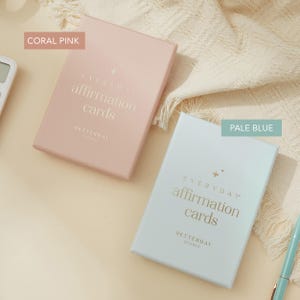 May include: Two boxes of affirmation cards in coral pink and pale blue. The boxes have gold text that reads "EVERYDAY affirmation cards" and "BETTERDAY STUDIO". The boxes are rectangular and are on a cream-colored surface.