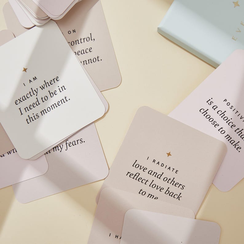 May include: A collection of square affirmation cards in shades of white, beige, and pink. Each card features inspirational text in a simple font, with a small star graphic. The cards are scattered on a light beige surface.