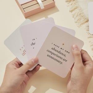 May include: A set of affirmation cards with rounded corners, held in hands. The cards are light pink and white, with text in a simple font. One card reads "I have my own abundance, comparisons are unnecessary."