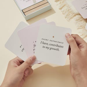 May include: A person holding a stack of square cards with inspirational text. The cards are white with a light pink gradient and gold star accents. The text on the top card reads "EVERY EXPERIENCE I have, contributes to my growth."