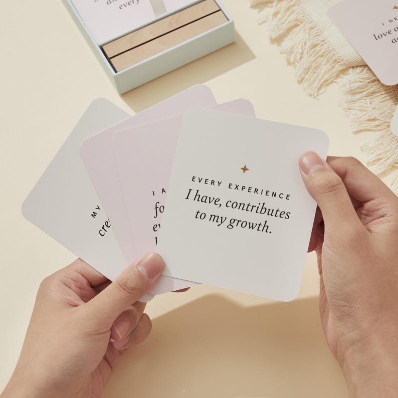May include: A person holding a stack of square cards with inspirational text. The cards are white with a light pink gradient and gold star accents. The text on the top card reads "EVERY EXPERIENCE I have, contributes to my growth."