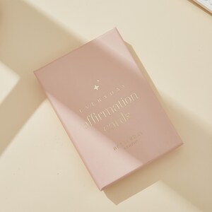 May include: A blush-colored box of "Everyday Affirmation Cards" with gold lettering and a star graphic. The box is rectangular and slightly angled, with the words "BETTERDAY STUDIO" at the bottom.