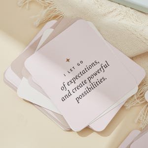 May include: A stack of square cards in shades of pink, beige, and white. The top card has the text "I LET GO of expectations, and create powerful possibilities." A small gold star is above the text.