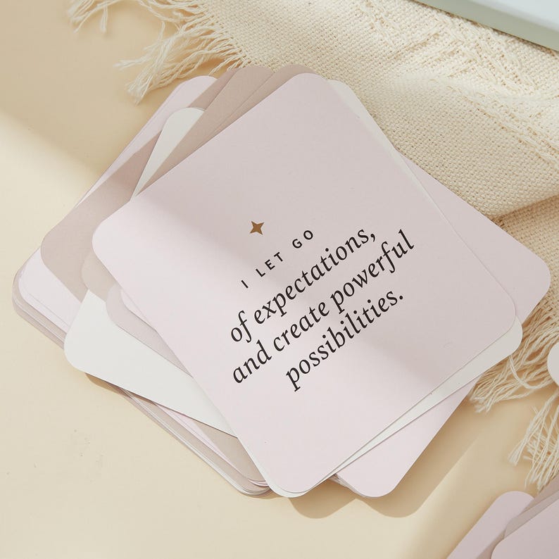 May include: A stack of square cards in shades of pink, beige, and white. The top card has the text "I LET GO of expectations, and create powerful possibilities." A small gold star is above the text.