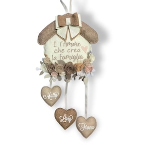 Può includere: Una casa in feltro bianco con un tetto in feltro marrone e un fiocco marrone. La casa ha un cuore rosa e il testo "E' l'amore che crea la famiglia". Ci sono tre cuori in feltro marrone appesi alla casa con i nomi "Matty", "Lory" e "Fiocco" scritti su di essi.