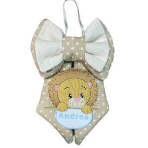 Può includere: Un appendiporta in tessuto beige e bianco con un fiocco, un viso di leone con un ovale blu con il nome "Andrea" su di esso e pois bianchi.