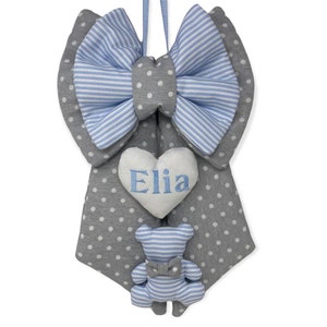 Può includere: Un portapacchi per porta per bambini grigio e blu con un grande fiocco, un cuore ricamato con il nome "Elia" e un piccolo orsacchiotto di peluche a righe.