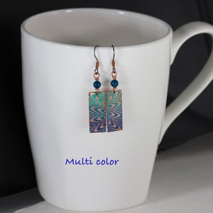 May include: A pair of earrings with a blue and green geometric design on a copper background. The earrings are hanging from a white mug. The earrings have a blue bead at the top of each earring.
