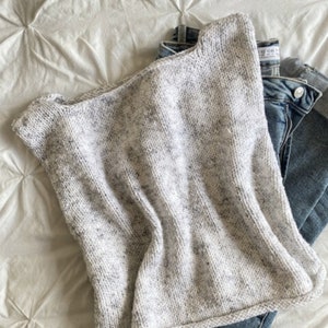 May include: A sleeveless, light gray knit top with a speckled pattern. The top is folded over a pair of blue denim jeans, resting on a white, quilted surface. The knitwear has a rounded neckline and a simple, casual design.