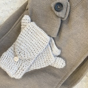 Puede incluir: Un par de guantes sin dedos de ganchillo beige con un detalle de botón. Los guantes están sobre un fondo de tela marrón.