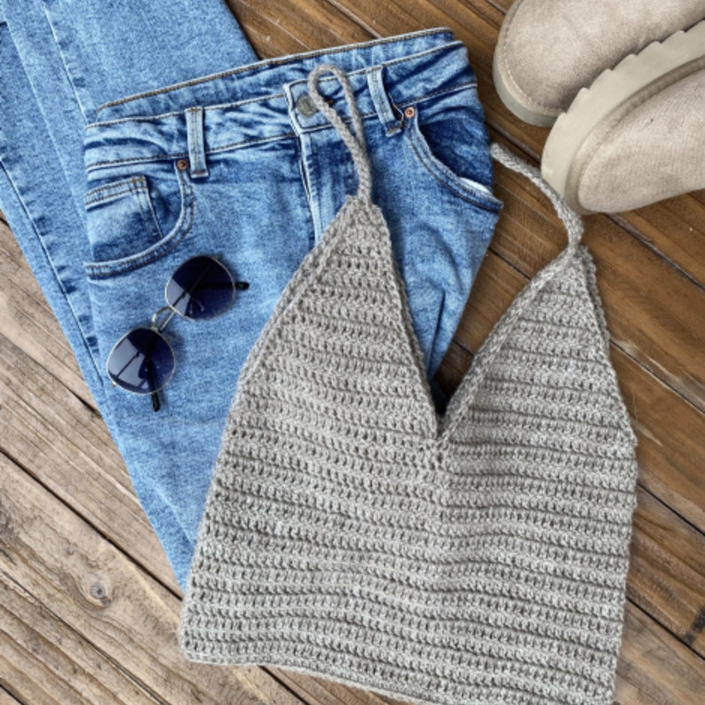 May include: A gray crocheted halter top with a V-neckline. The top is laying on a wooden surface with a pair of blue jeans and sunglasses.