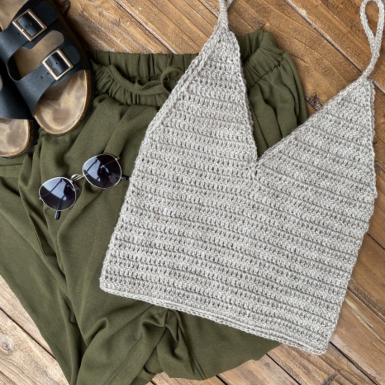 May include: A beige crochet tank top with a V-neck and thin straps. The top is laying on a green fabric background.