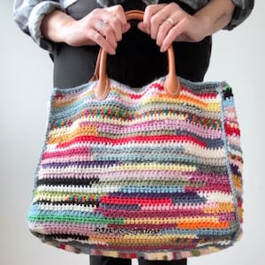 May include: A large, colourful crochet tote bag with a rectangular shape. The bag features a vibrant patchwork design with horizontal stripes in various colours, including red, blue, yellow, and purple. It has brown leather handles.