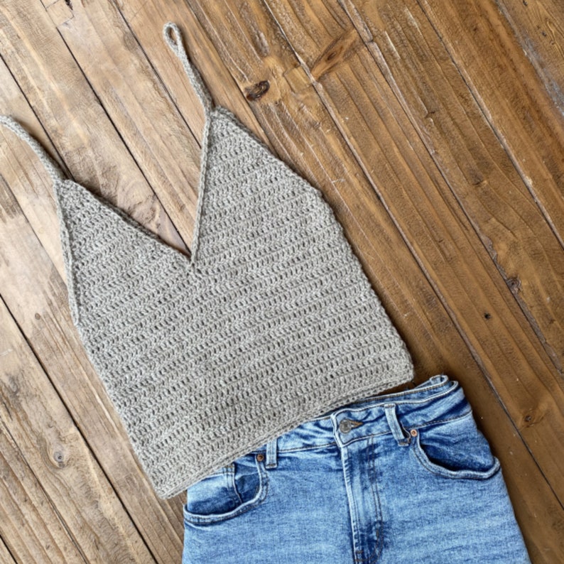 May include: A gray crochet tank top with a v-neck and thin straps, laying on top of a pair of blue denim jeans.