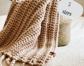 May include: A crocheted throw blanket in a light brown colour with fringe on the bottom edge. The blanket is folded and draped over a wicker basket. The text "5 sizes" is written on a white circle.