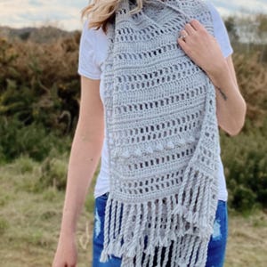 May include: A grey crochet scarf with a lacy pattern and fringe. The scarf is draped around a person's neck.
