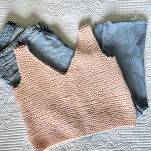 May include: A light peach-colored knitted tank top with a V-neck design. The top is paired with light blue denim jeans. The items are arranged on a white, textured surface, suggesting a casual outfit.