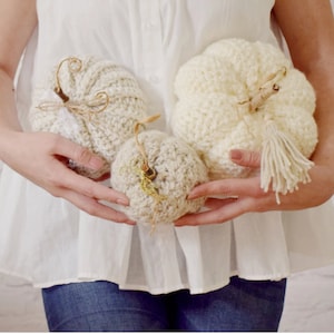 May include: Three crocheted pumpkins in white and cream colors. The pumpkins are decorated with yarn tassels and are held in the hands of a person.
