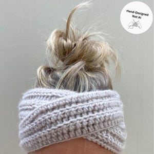 May include: A light gray crochet ear warmer headband featuring a twisted front design with a textured ribbed stitch pattern. A circular white badge in the upper right corner displays the text Hand-Designed Not AI with a line drawing of hands knitting.