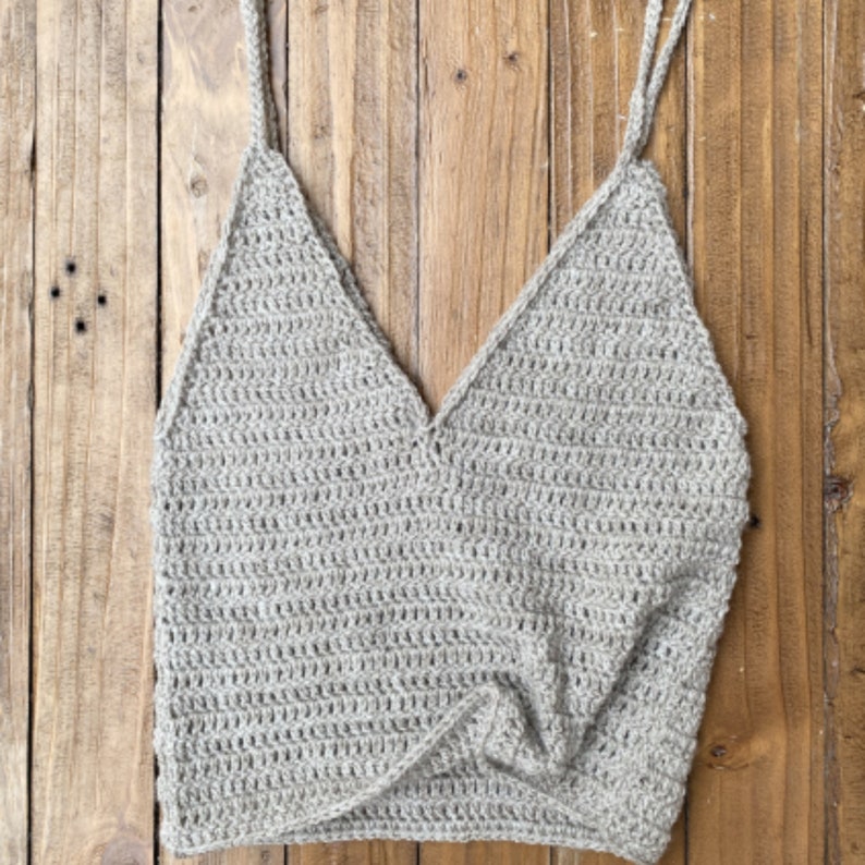 May include: A beige crochet tank top with a V-neck and thin straps. The top is made with a simple stitch pattern and has a knotted detail at the front.
