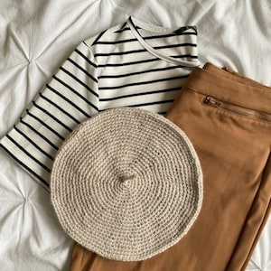 Peut inclure: Un béret en crochet beige, un chemisier rayé noir et blanc et un pantalon marron avec une fermeture éclair.