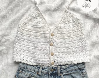 Op de afbeelding: Een wit mouwloos gehaakt crop topje met een V-hals, dunne schouderbandjes en een knoopsluiting aan de voorzijde met vier lichtbruine knopen. De top is gecombineerd met een lichtblauwe spijkerbroek. Een sticker vermeldt Hand-Designed Not AI.