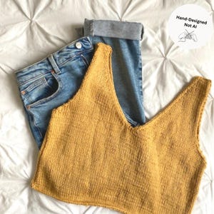 May include: A mustard yellow sleeveless knit tank top with a deep V-neckline paired with blue denim jeans featuring a rolled cuff. A circular white graphic in the corner contains the text Hand-Designed Not AI with an illustration of hands knitting.