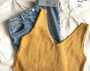 May include: A mustard yellow sleeveless knit tank top with a deep V-neckline paired with blue denim jeans featuring a rolled cuff. A circular white graphic in the corner contains the text Hand-Designed Not AI with an illustration of hands knitting.