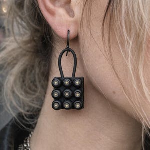 Könnte beinhalten: Schwarze Metallohrringe mit einem einzigartigen Design. Jeder Ohrring hat eine rechteckige Form mit neun kleinen, runden, goldfarbenen Akzenten. Die Ohrringe hängen an einem gebogenen, länglichen Haken. Der Schmuck wird von einer Person getragen.
