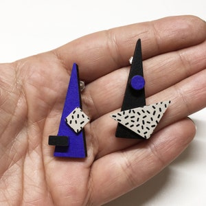 May include: Geometric stud earrings with a modern design. One earring is a blue triangle with a black square and a white and black square. The other earring is a black triangle with a blue circle and a white triangle with black speckles.