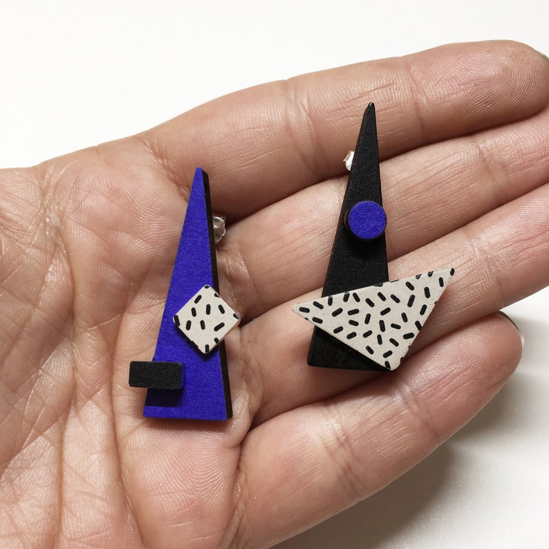May include: Geometric stud earrings with a modern design. One earring is a blue triangle with a black square and a white and black square. The other earring is a black triangle with a blue circle and a white triangle with black speckles.