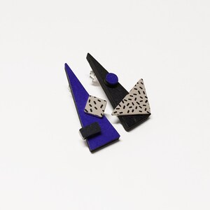 May include: Geometric earrings featuring a bold design. One earring is a long, blue triangle with a black rectangle and a square with a speckled pattern. The other earring is a black triangle with a speckled triangle and a blue circle.