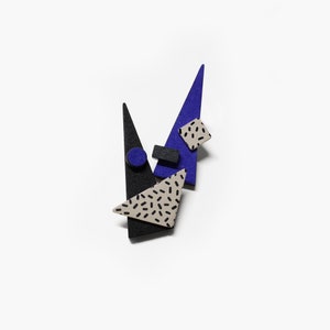 May include: An abstract brooch featuring geometric shapes in black, blue, and beige. The design includes triangles, a square, a rectangle, and a circle, with a speckled pattern on some elements. The brooch is set against a white background.