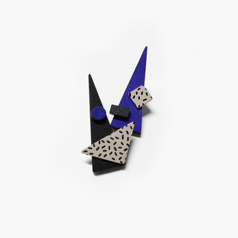 May include: An abstract brooch featuring geometric shapes in black, blue, and beige. The design includes triangles, a square, a rectangle, and a circle, with a speckled pattern on some elements. The brooch is set against a white background.