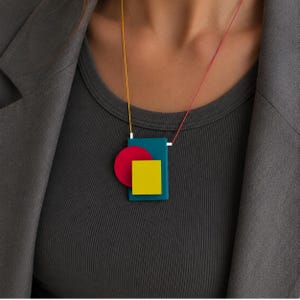 Peut inclure: Un collier pendentif g&eacute;om&eacute;trique avec un rectangle turquoise, un cercle rouge et un carr&eacute; jaune. Le pendentif est suspendu &agrave; un cordon jaune et rouge. Le collier est port&eacute; sur un haut et un blazer gris.