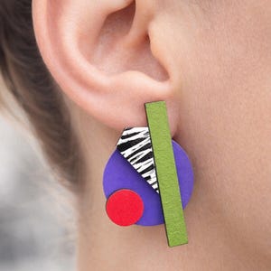Pode incluir: Brincos geométricos abstratos. Apresentam um círculo roxo, um círculo vermelho, um retângulo verde e um triângulo com estampa de zebra em preto e branco. Design moderno e colorido.