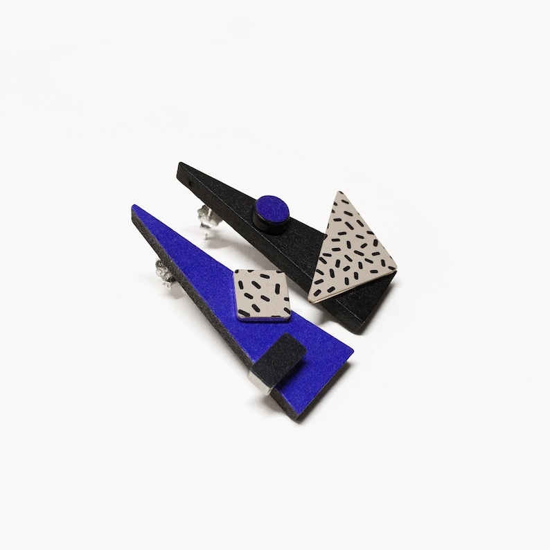 May include: Geometric stud earrings in black, blue, and silver. The earrings feature a triangular shape with a blue felt-like surface, a black triangle, and a silver triangle with black speckles. A small black square and circle are also included.