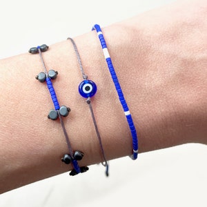 Puede incluir: Tres pulseras delicadas en tonos azules y grises. Una presenta cuentas azules y detalles grises, otra tiene un dije de ojo turco azul, y la tercera es una cadena de cuentas azules con detalles blancos. Estas pulseras son perfectas para el uso diario.