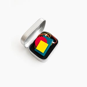Geometric Bauhaus style statement necklace stored in a minimalist metal gift tin, colorful abstract wooden pendant jewelry with Memphis inspired design, modern avant garde necklace packaging, artistic contemporary fashion accessory on soft neutral background