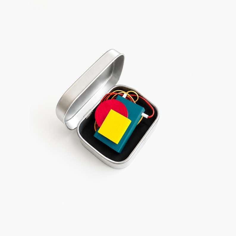 Geometric Bauhaus style statement necklace stored in a minimalist metal gift tin, colorful abstract wooden pendant jewelry with Memphis inspired design, modern avant garde necklace packaging, artistic contemporary fashion accessory on soft neutral background